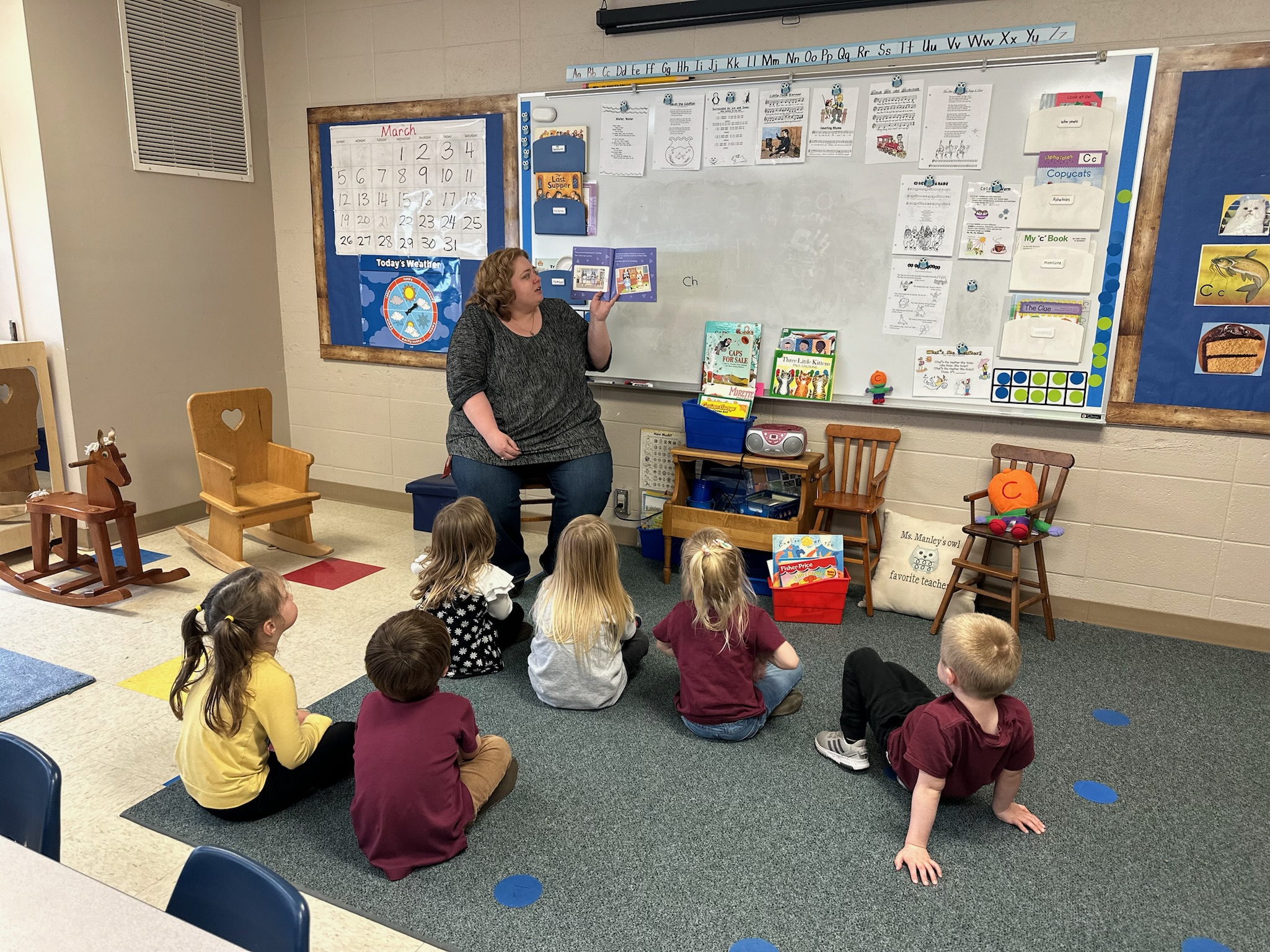 A female teacher sits in a wooden chair and holds up the orange pages of a book as she reads to her preschool students who are sitting on the blue carpet in front of her. 