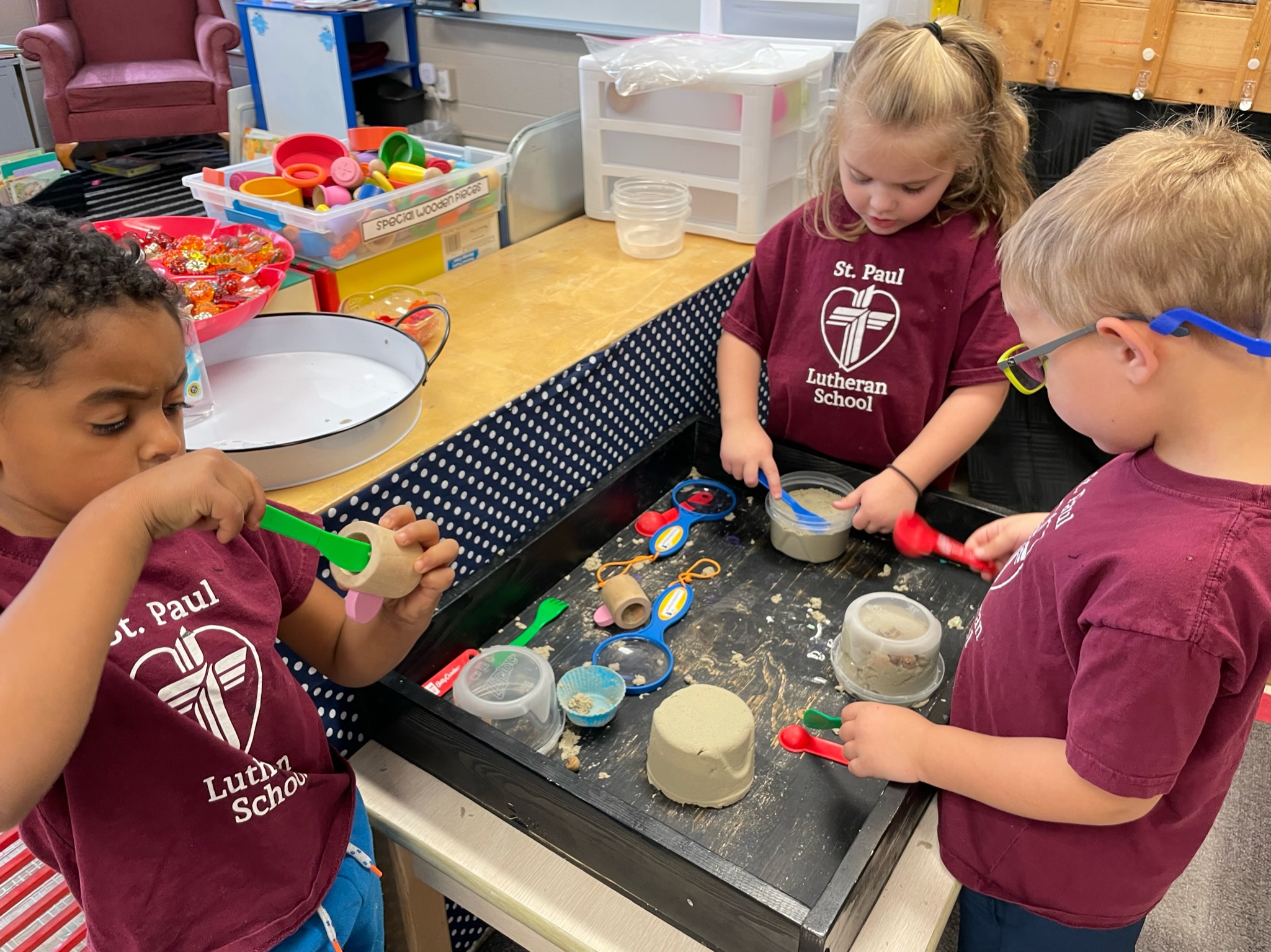 A caucasian boy with glasses, a caucasian girl, and an African American boy all wearing maroon colored uniform t-shirts are playing in a sand sensory bin in their preschool classroom. 