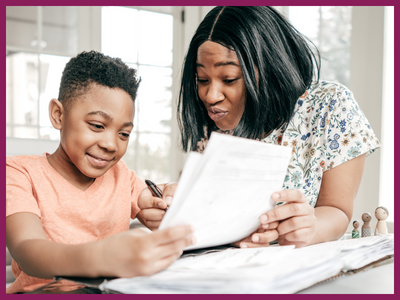 An African-American mother and son sit at a table together looking at homework. The mom is making a silly face.