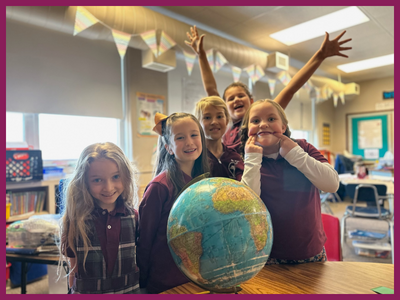 Six St. Paul Lutheran School elementary-age female students smile as they stand behind a globe. One has her fingers on the corners of her mouth to make a silly face. The girl in the back has thrown her arms into the air as she cheers.
