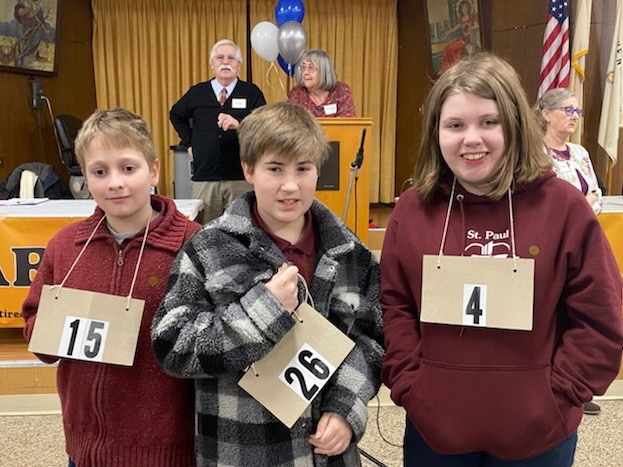 Three middle school students wearing cardboard numbers around their neck pose for a photo before their spelling bee contest. 