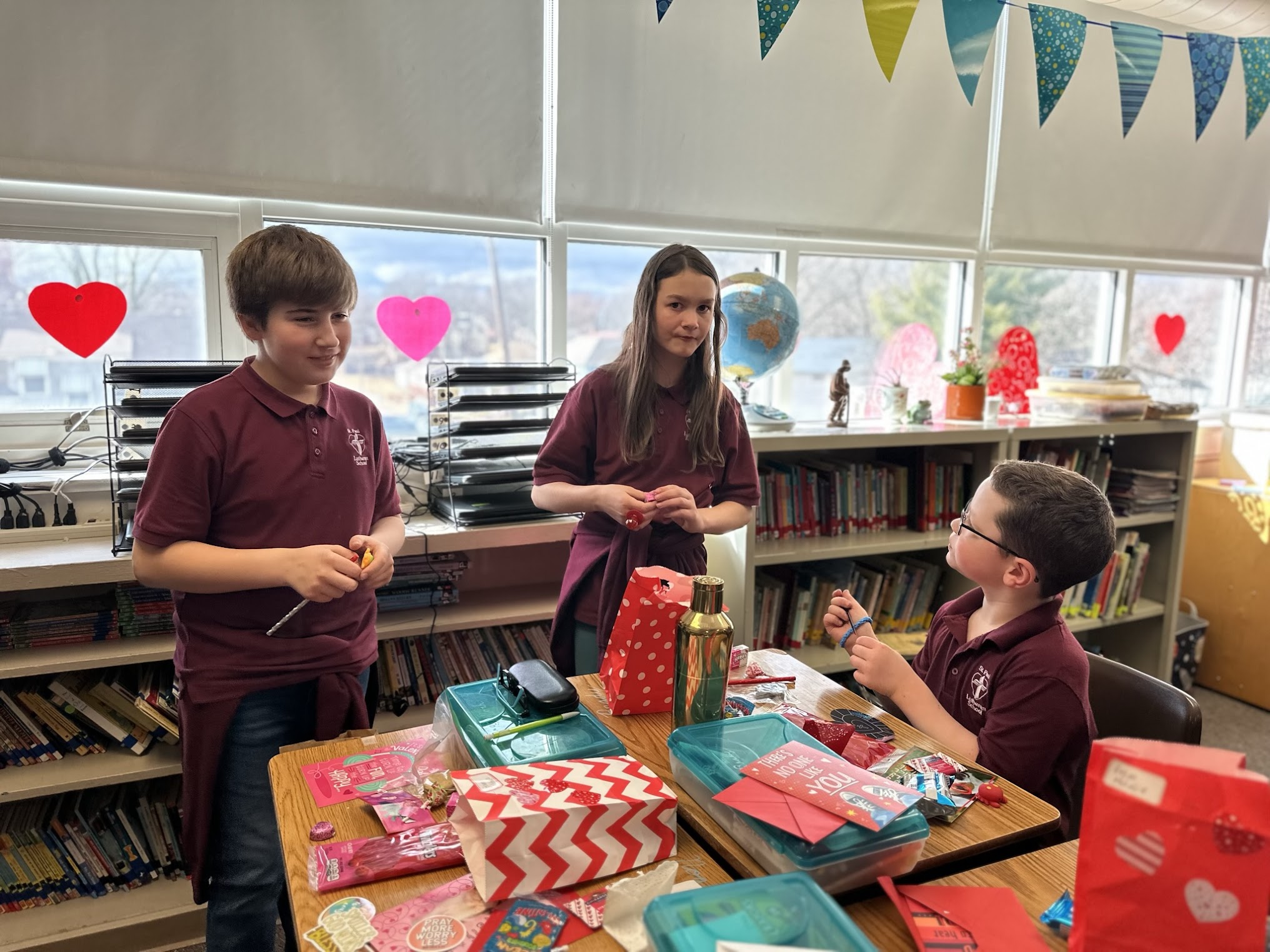 Three fifth grade students prepare for a Valentine's Day party. One caucasian boy and one caucasian girl are standing. A caucasian boy is seating at his desk and looking up at them. 